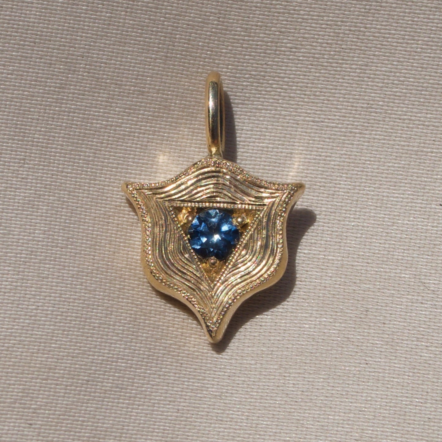 A gold shield-shaped pendant with a textured, wavy pattern and a central cluster of blue gemstones, displayed on a neutral beige textured fabric background.