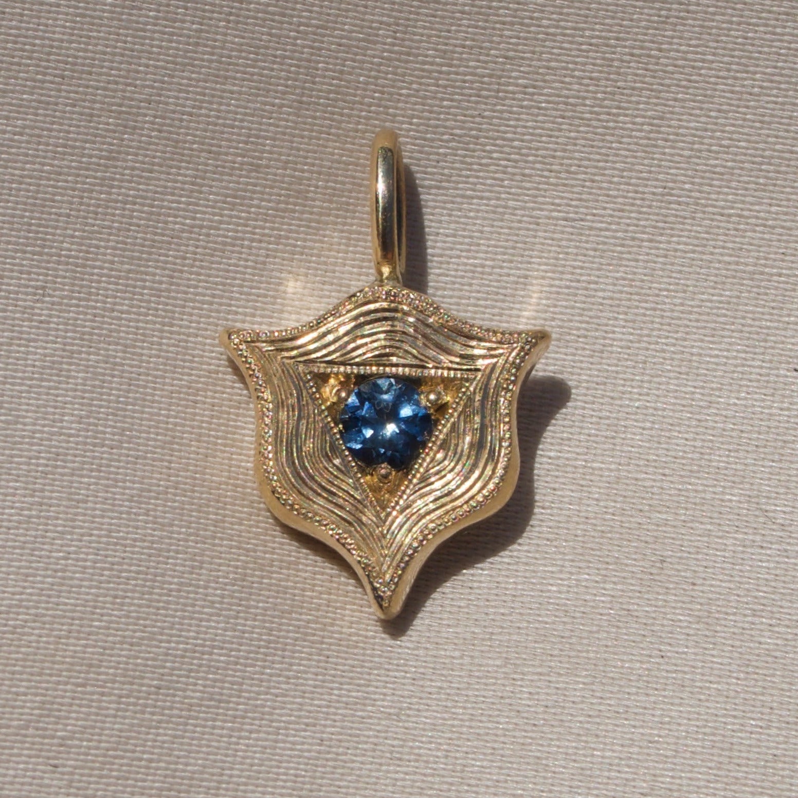 A gold shield-shaped pendant with a textured, wavy pattern and a central cluster of blue gemstones, displayed on a neutral beige textured fabric background.