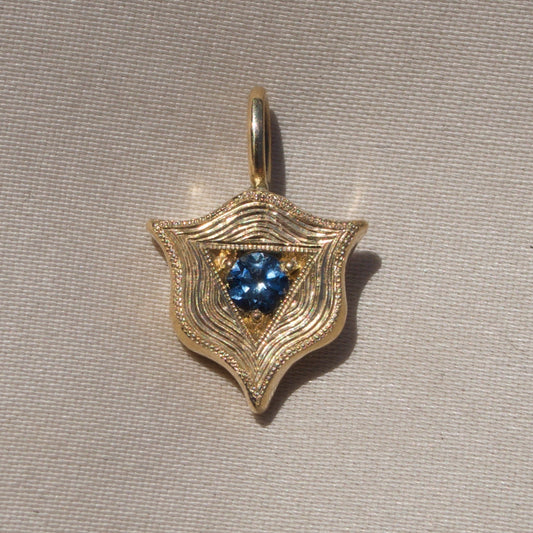 A gold shield-shaped pendant with a textured, wavy pattern and a central cluster of blue gemstones, displayed on a neutral beige textured fabric background.