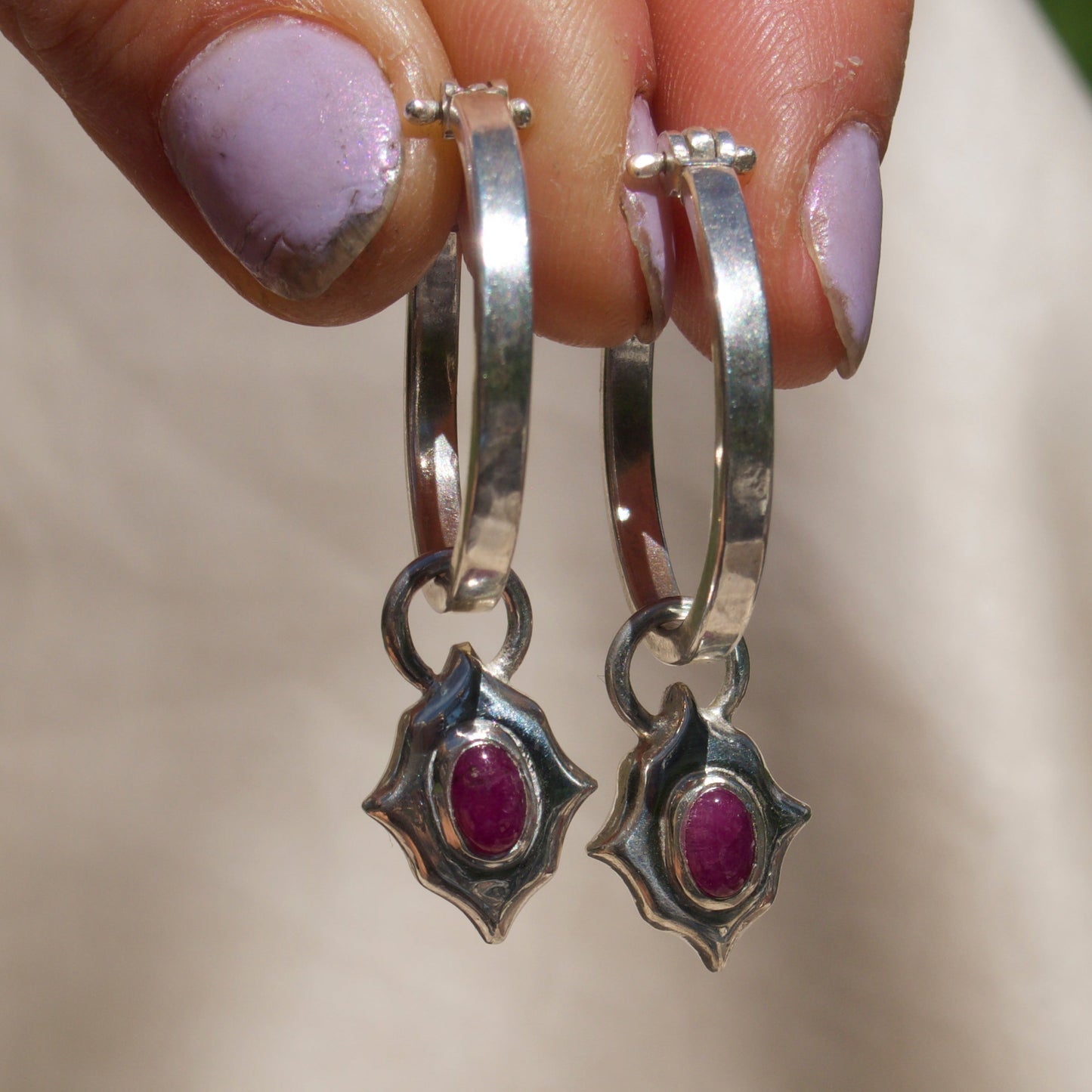 A close-up of a hand holding a pair of silver hoop earrings with starburst charms set with oval pink gemstones, against a soft beige fabric background.